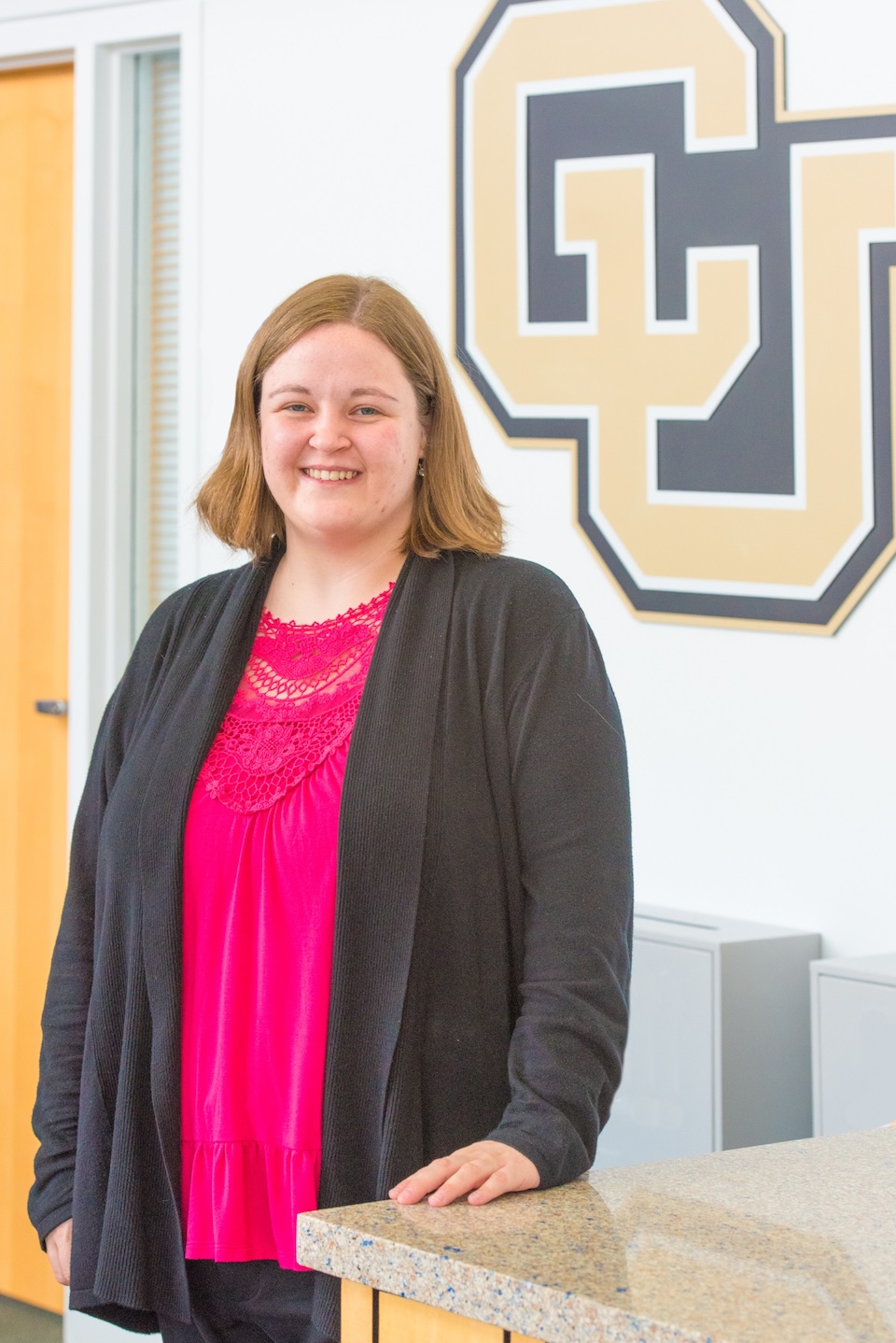 Alyssa has medium length dark blonde hair and is wearing a pink shirt under a dark gray cardigan and is standing in front of a CU logo