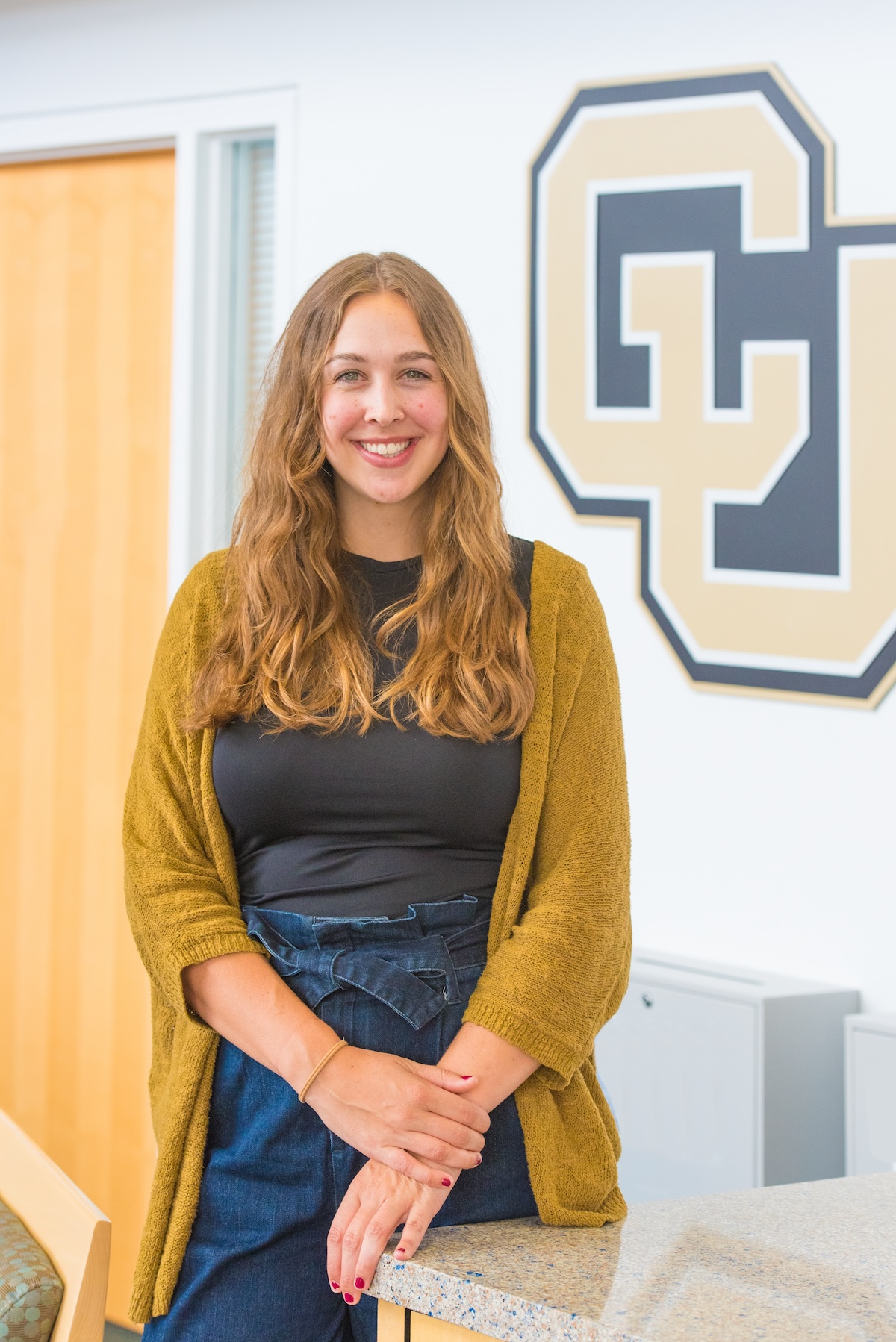 Kaelyn has long dark blonde hair and is wearing a black shirt under a gold cardigan and is standing in front of a CU logo