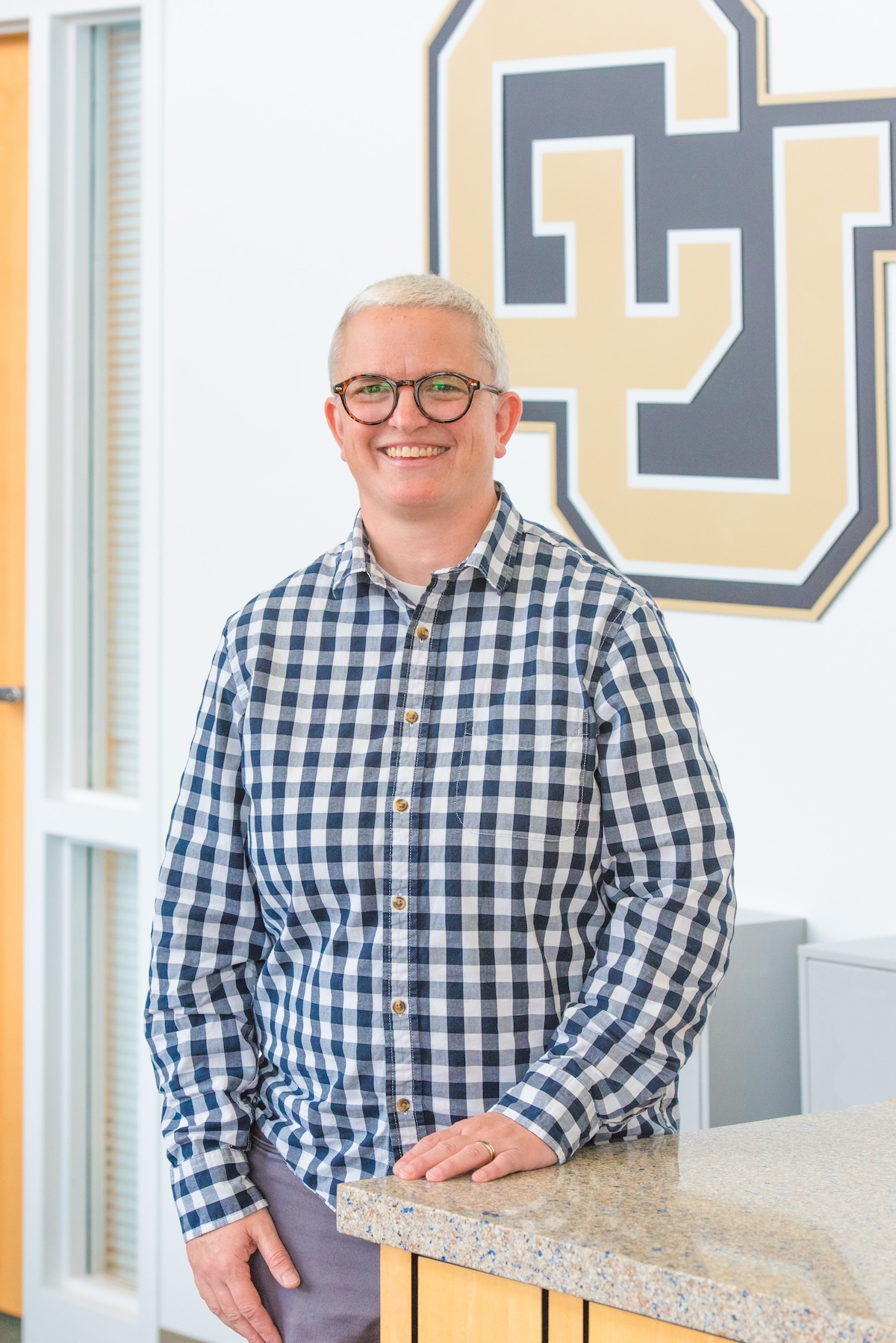 Lauren has short gray hair and glasses and is wearing a blue and white checked shirt standing in front of a CU logo