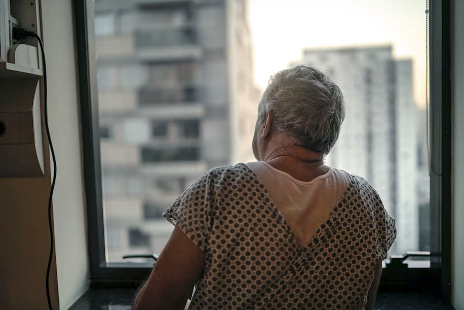 Male senior citizen in hospital gown looking out window