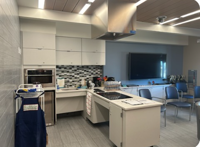 The Cardiac Kitchen: white cabinets aith a white, black and grey glass tile backsplash stands next to a room with blue chairs and a TV mounted to a wall.