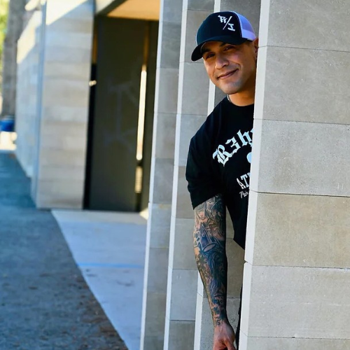 SOC Hero- Tattooing Hope May 2025 Mario peeks out from behind a concrete wall with a smile on his face. He is wearing a baseball cap and his arm is covered in artistic tattoos.