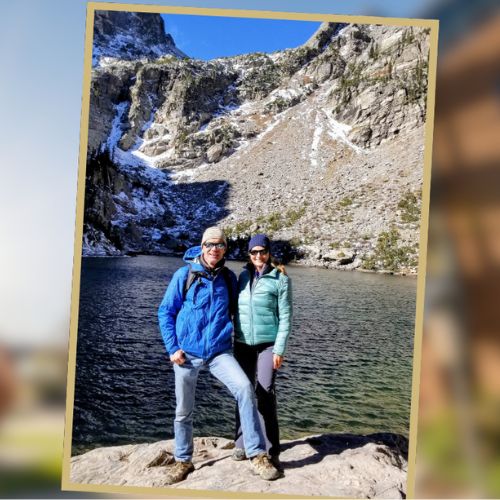 Jim, in a blue coat and jeans, poses for a photo with his wife Anita, in a teal jacket, on a hike near a high altitude lake in the Rocky Mountains.