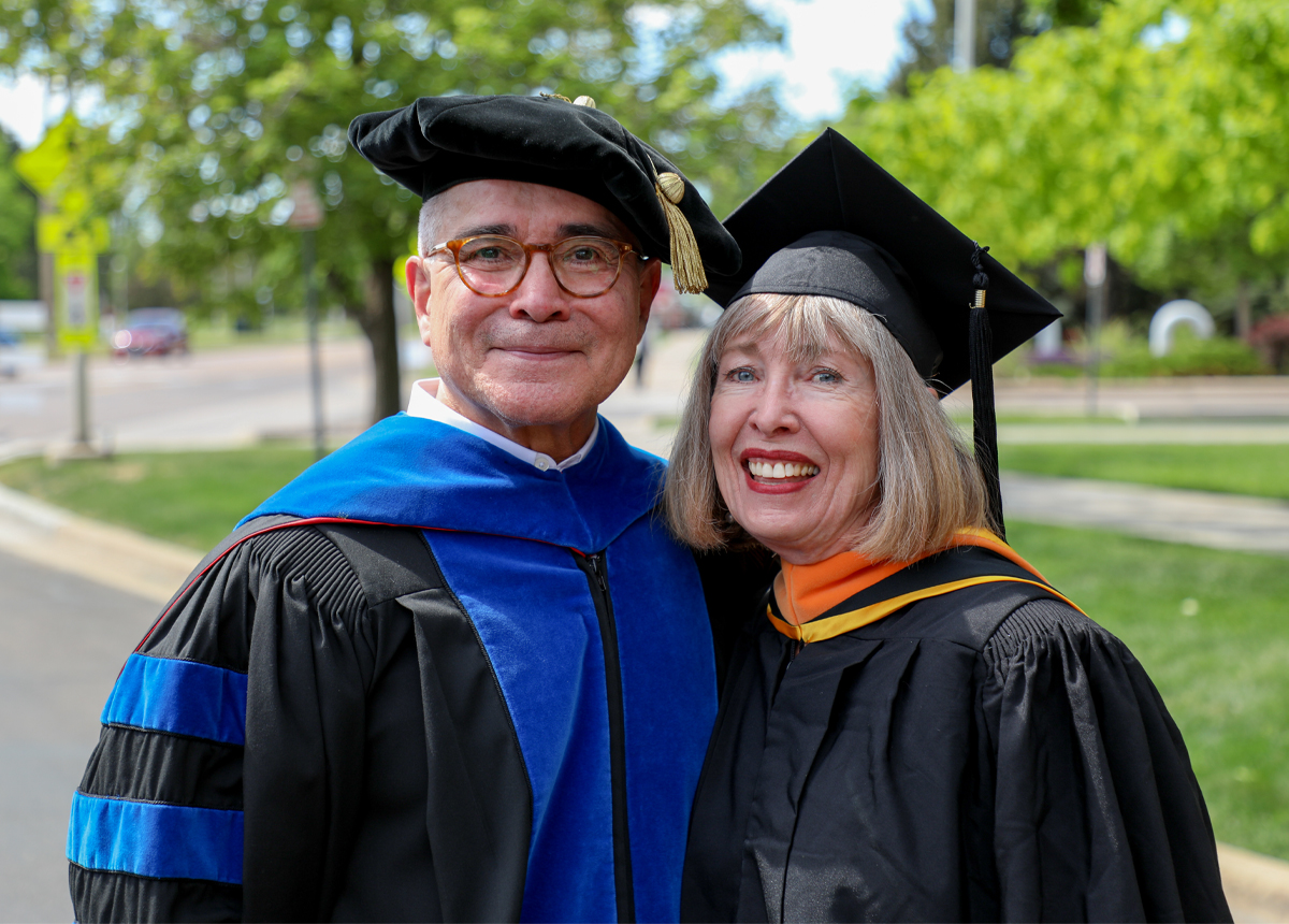 Dean Eli and Karen Zink pose for a photo together wearing ceremonial graduation caps and gowns.
