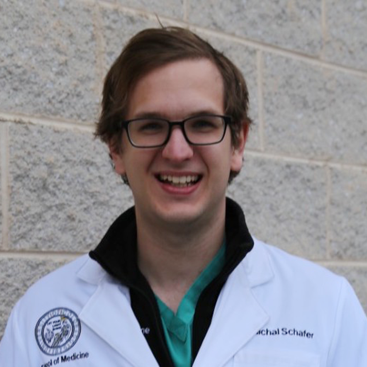 Michal Schäfer, medical scholar. He is smiling at the camera. He is wearing a white medical coat and standing outdoors in front of a gray brick wall.