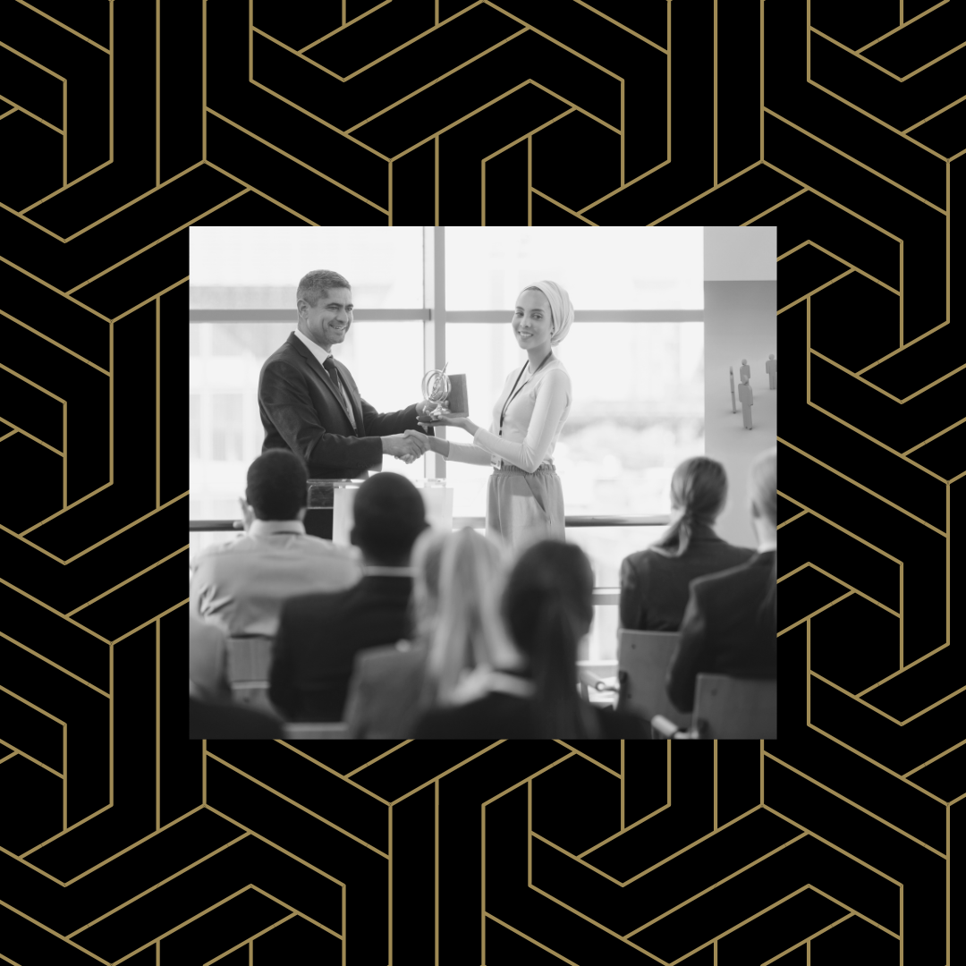 Black and white image of someone handing another person an award in front of an audience over a black and gold geometric background.