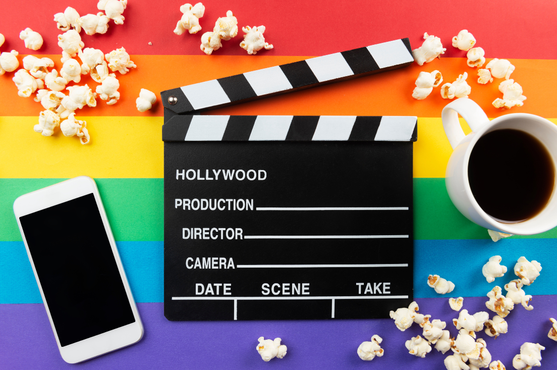 Image of a clapboard, cell phone, cup of coffee, and popcorn sitting on top of a rainbow flag