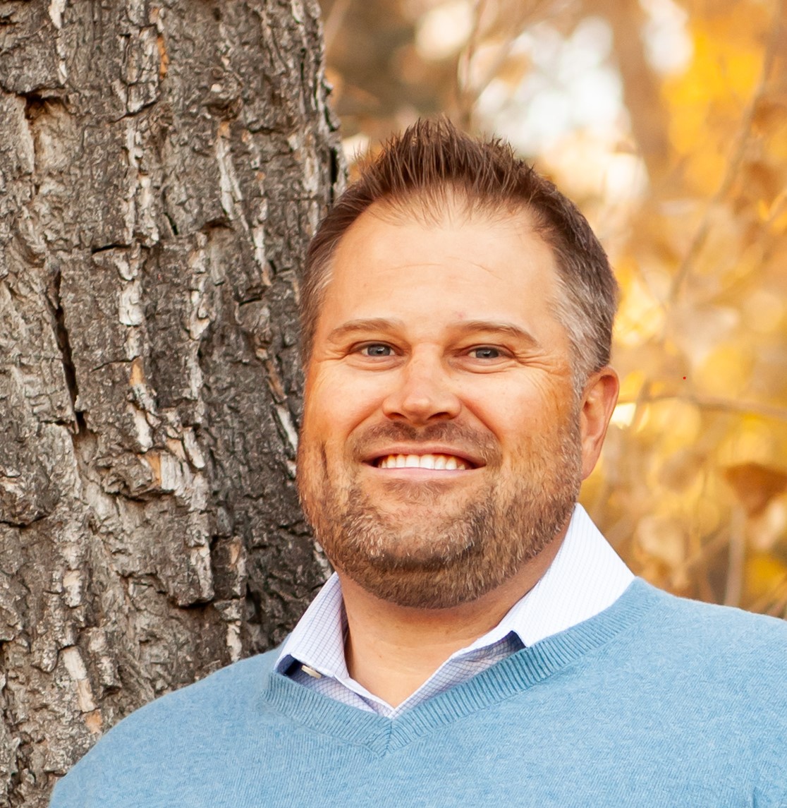 picture of a mid-40s White bearded male in a light blue sweater next to a tree
