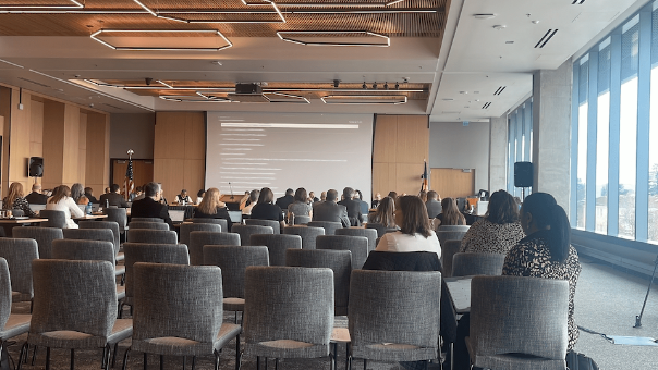 Picture of the back of a meeting while attendees look at a presentation on a screen with OIT AV support managing it.